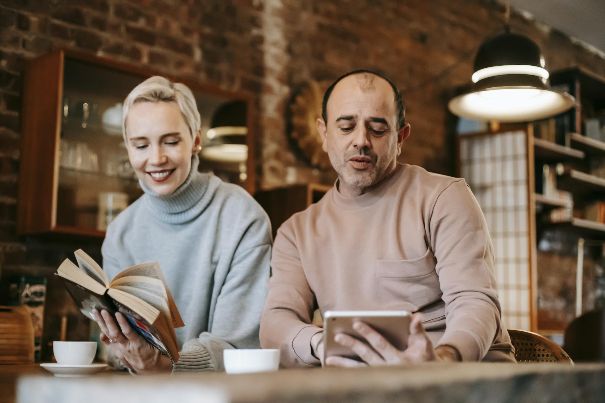 Adult multiracial spouses sitting at table while using tablet and reading book at hone