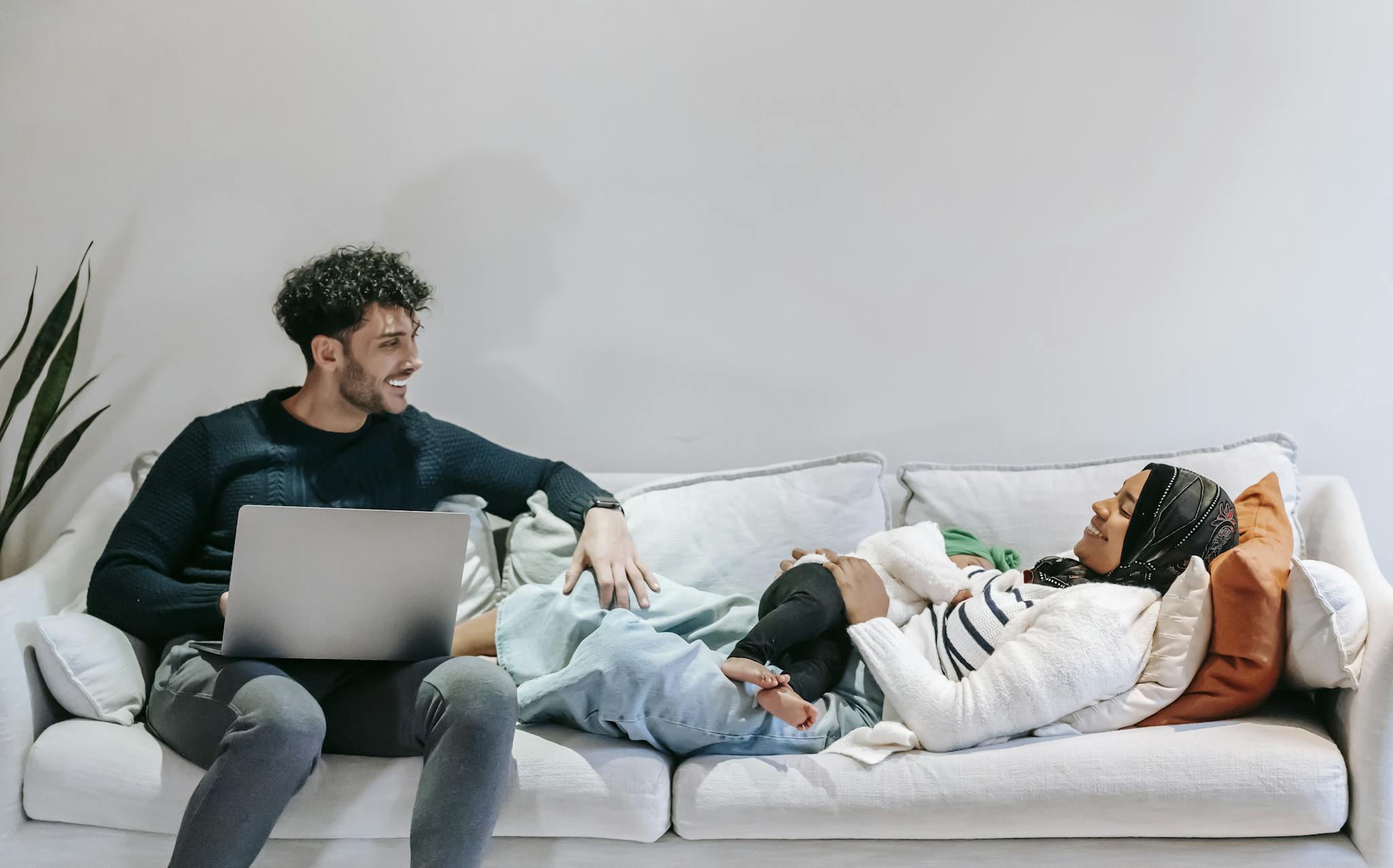 Cheerful young black Muslim woman in casual clothes and hijab relaxing on bed with cute infant in arms and communicating with ethnic husband working remotely on laptop