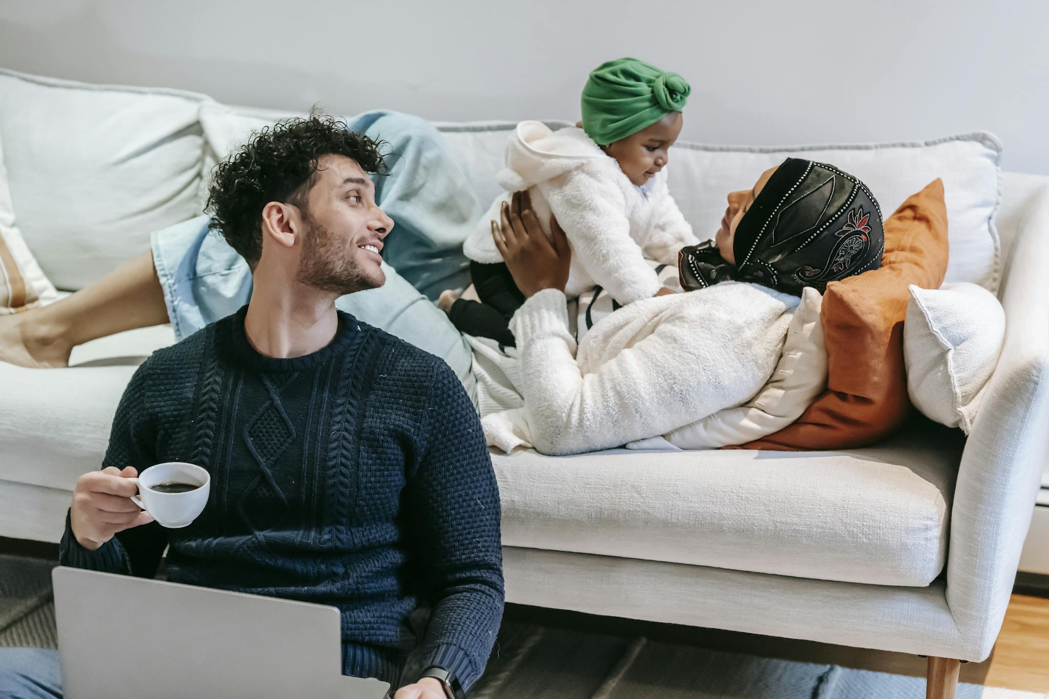 Happy young ethnic man in casual clothes drinking coffee and working on laptop while sitting on floor near Muslim wife lying on sofa and playing with cute toddler daughter